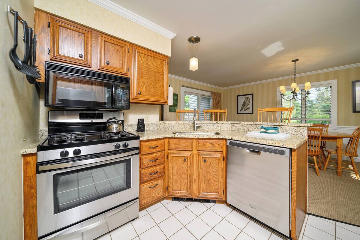 A kitchen with wooden cabinets, a stove, microwave, and a dishwasher. There is a dining area with chairs and a table.
