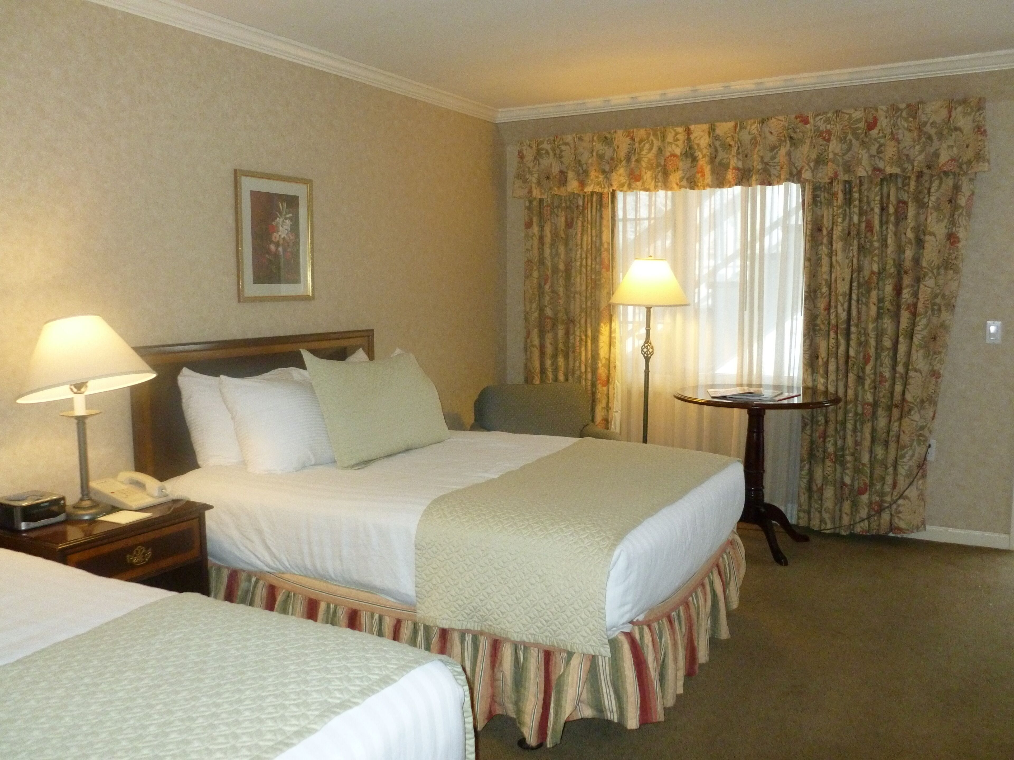 Habitación de hotel con dos camas, una mesita de noche con lámpara, una pequeña mesa con lámpara y una ventana con cortinas.