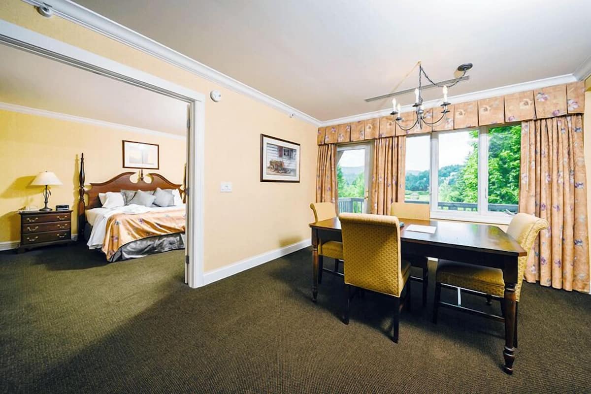 A hotel room with a bed, a dining table with chairs, a chandelier, and a window with curtains.