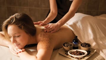 A person receiving a back massage on a bed with a tray of coffee beans and two small bowls of what appears to be massage oil.