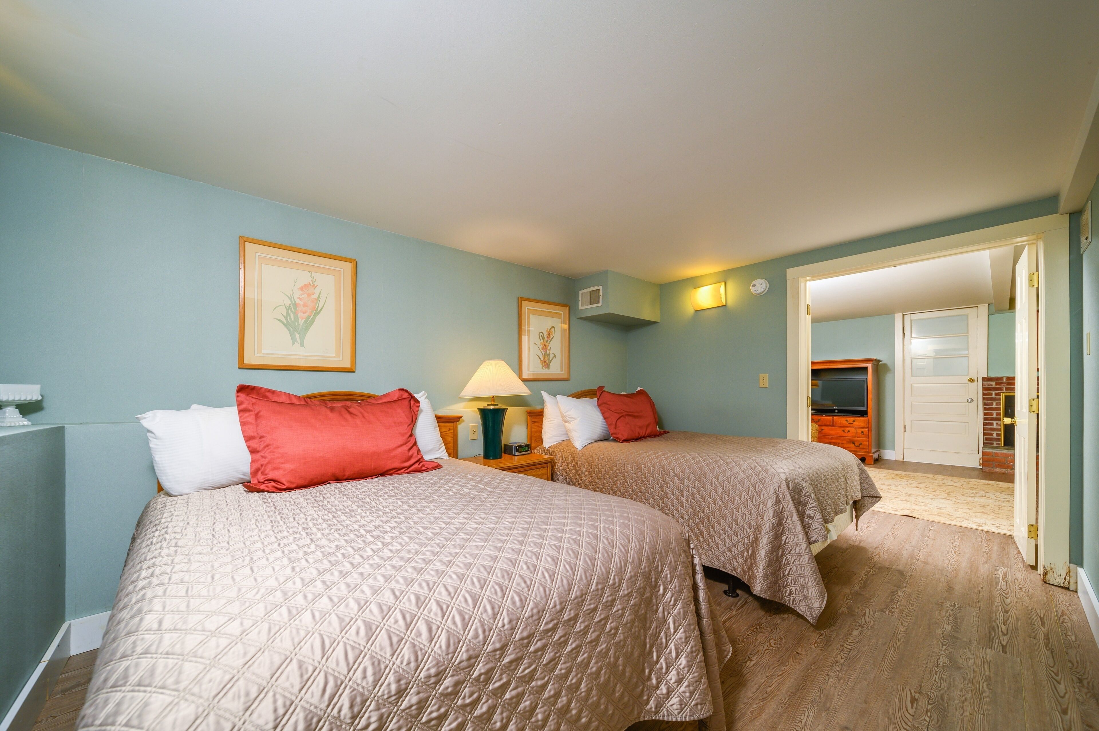 A hotel room with two beds, a nightstand with a lamp, and a door leading to another room.