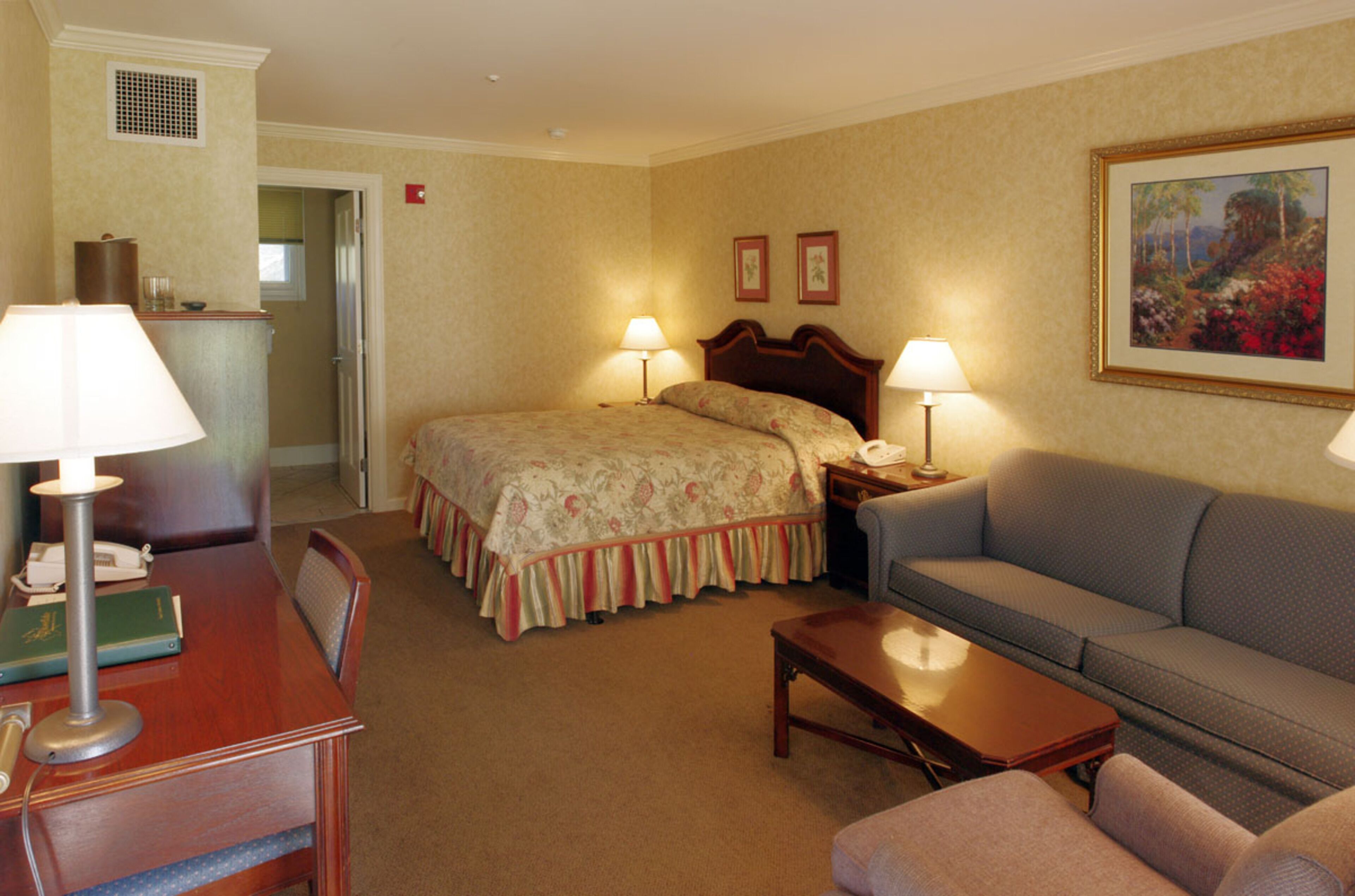 A hotel room with a bed, a sofa, a desk with a lamp, and a painting on the wall.