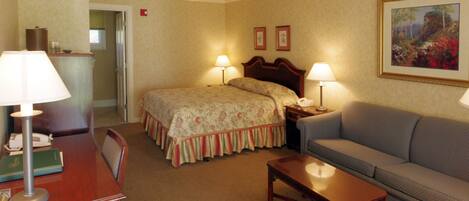 A hotel room with a bed, a sofa, a desk with a lamp, and a painting on the wall.