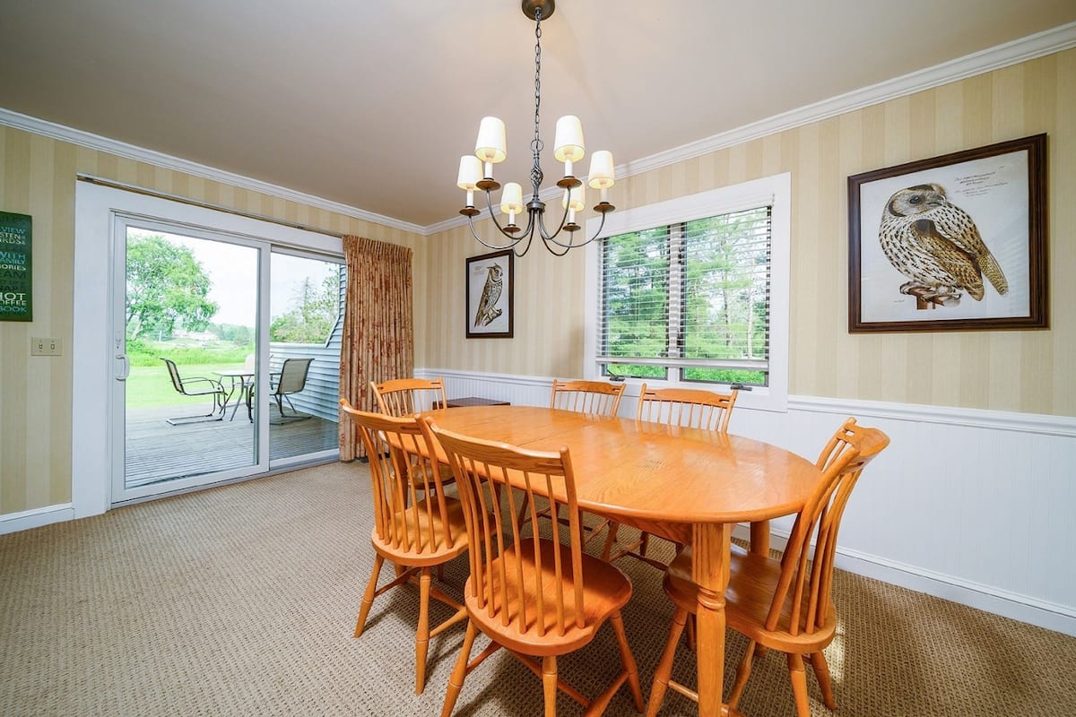 A dining room with a large wooden table and chairs, a chandelier, and two windows with blinds.