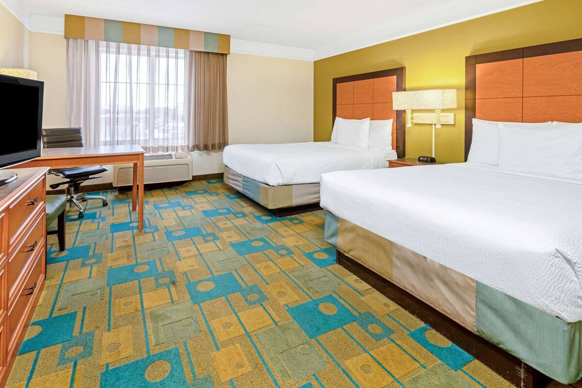 Quarto de hotel com duas camas, uma mesa, uma televisão e um tapete estampado.