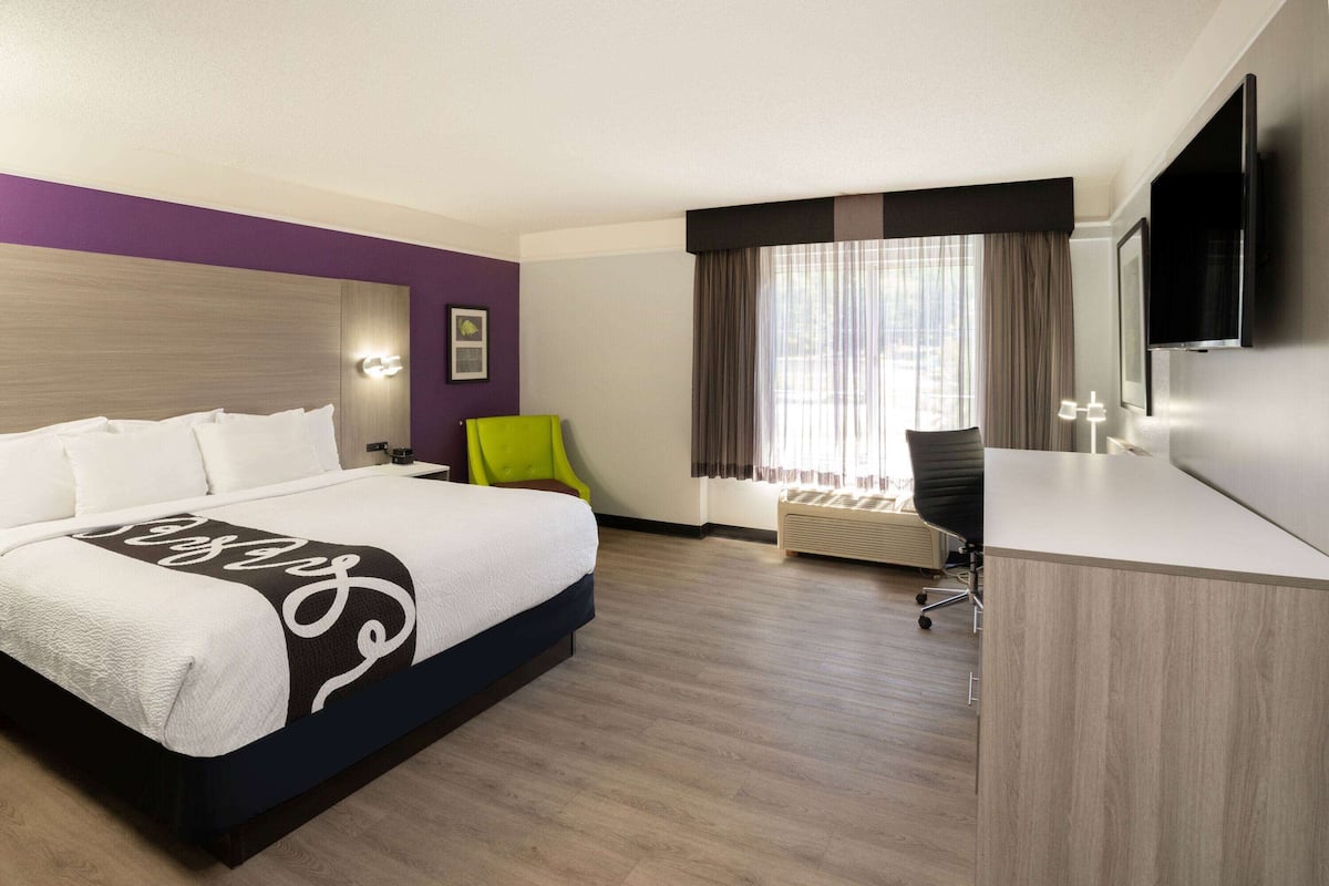 Habitación de hotel con cama, escritorio, silla, televisor y ventana con cortinas.