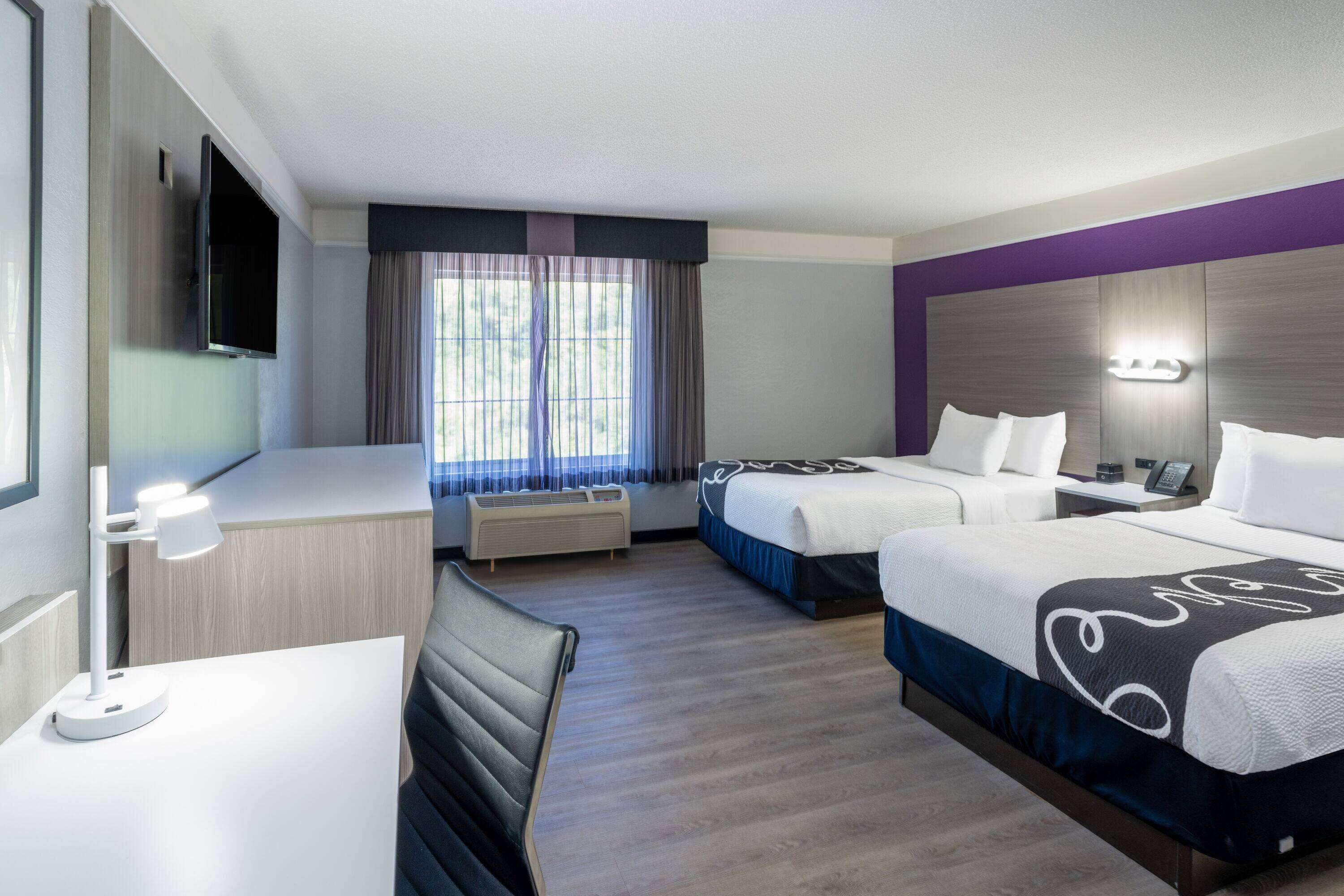 A hotel room with two beds, a desk, a TV, and a window with curtains.
