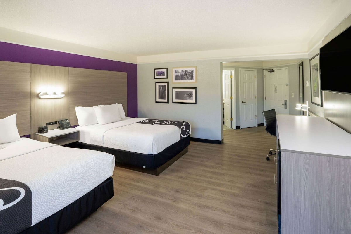 A hotel room with two beds, a desk, a mirror, and framed pictures on the wall.