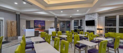 A modern dining area with multiple tables and chairs, a reception desk, and a TV screen.