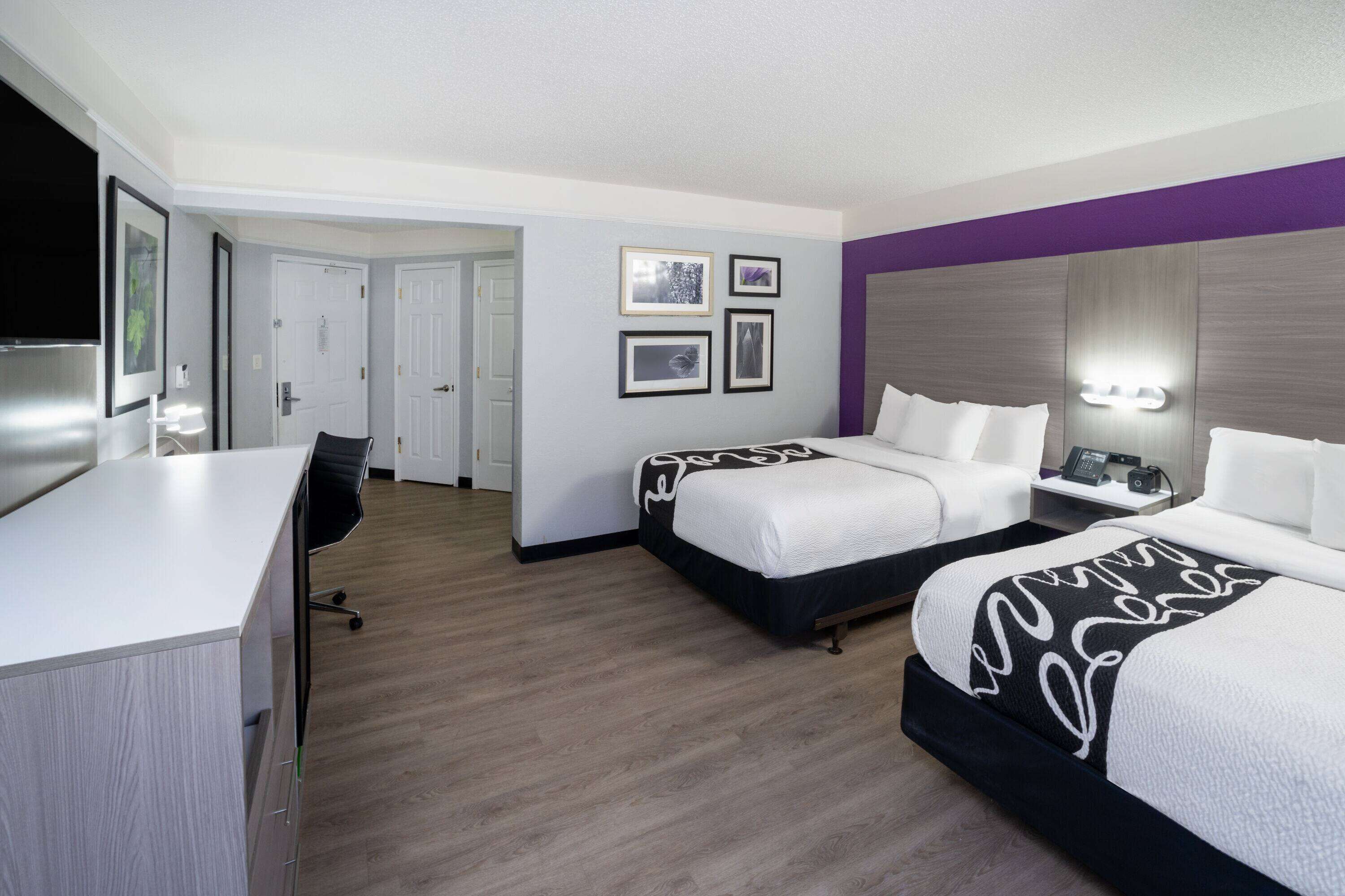 A hotel room with two beds, a desk, and a chair. There are framed pictures on the wall and a nightstand with a lamp.