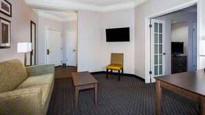 Habitación de hotel con sofá, una mesita, una silla, un televisor montado en la pared y una puerta que da a otra habitación.