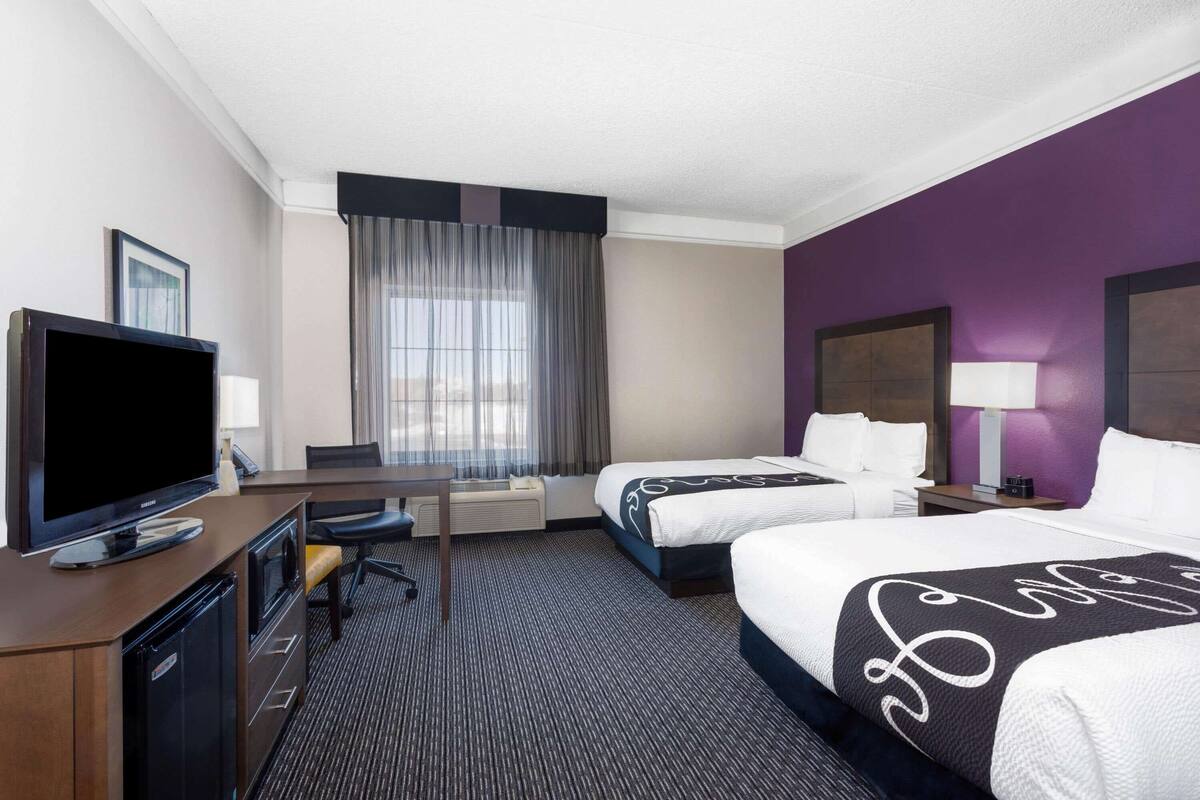 Ein Hotelzimmer mit zwei Betten, einem Schreibtisch mit Fernseher, einem Sessel, einem Fenster mit Vorhängen und einer Wand in Lila.