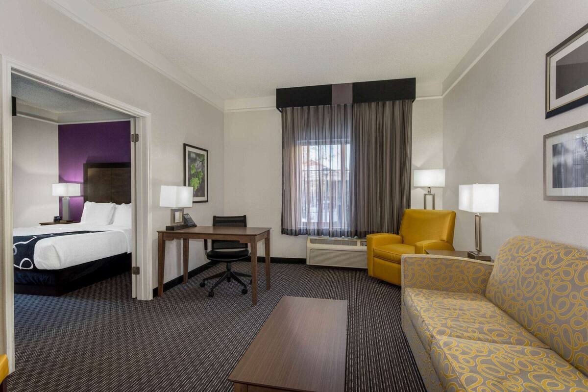 Ein Hotelzimmer mit Bett, Schreibtisch, gelbem Sessel, Sofa und einem Fenster mit Vorhängen.