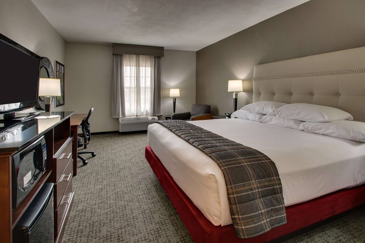 A hotel room with a large bed, a desk with a television, two lamps, and a window with curtains.