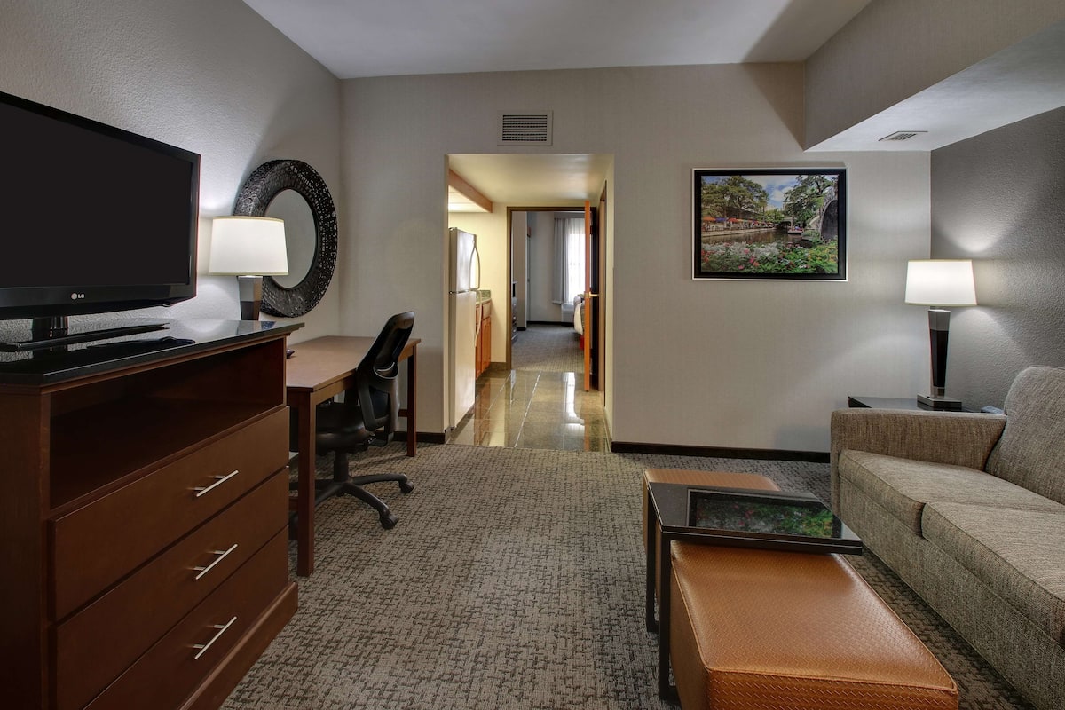A hotel room with a flat-screen TV, a desk with a chair, a sofa, a coffee table, a lamp, and a painting on the wall.