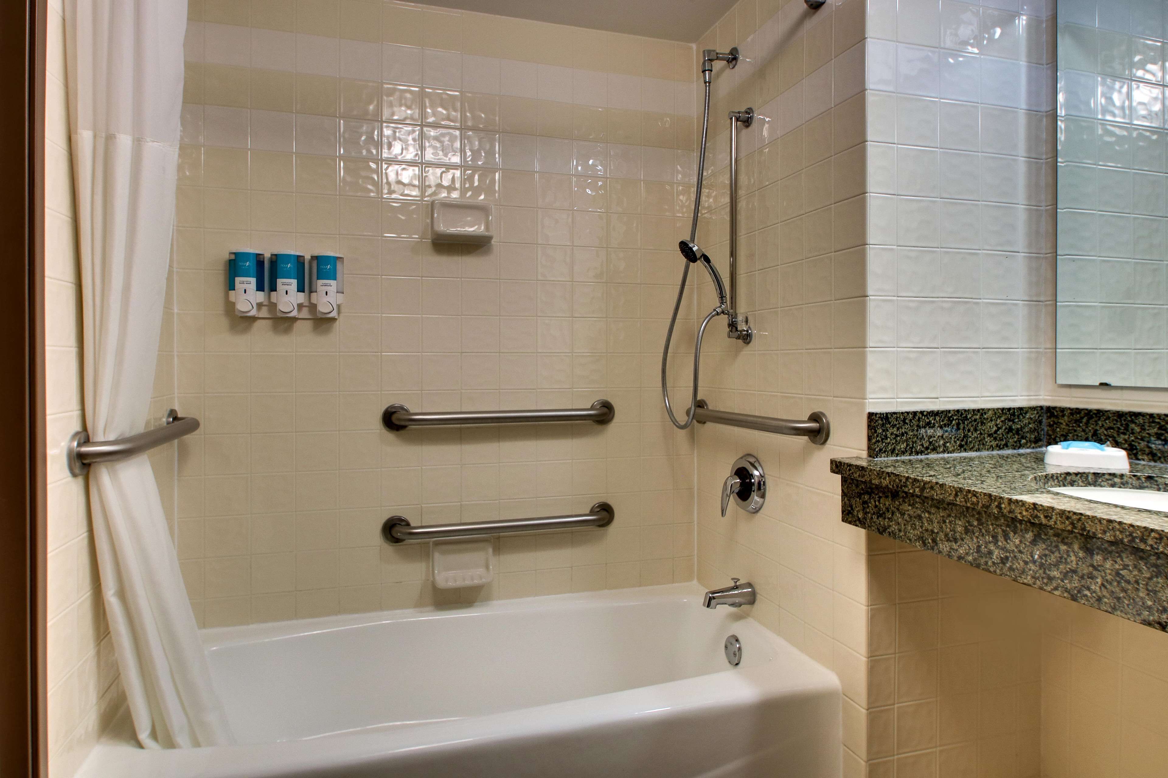 Un baño con bañera, barras de apoyo, cabezal de ducha y encimera con dispensador de jabón.