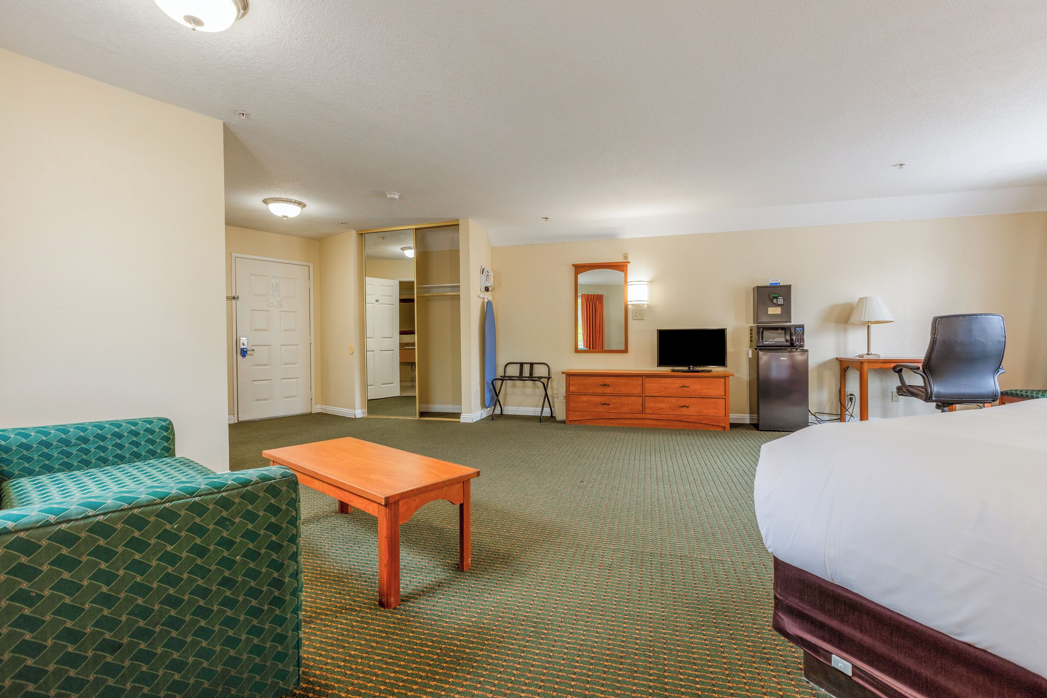 A hotel room with a bed, a green upholstered sofa, a wooden table, a television, a microwave, a desk, and a mirror.