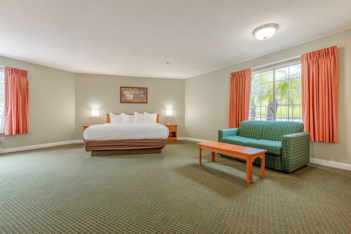 A hotel room with a bed, a green armchair, a wooden table, a nightstand, and a window with curtains.