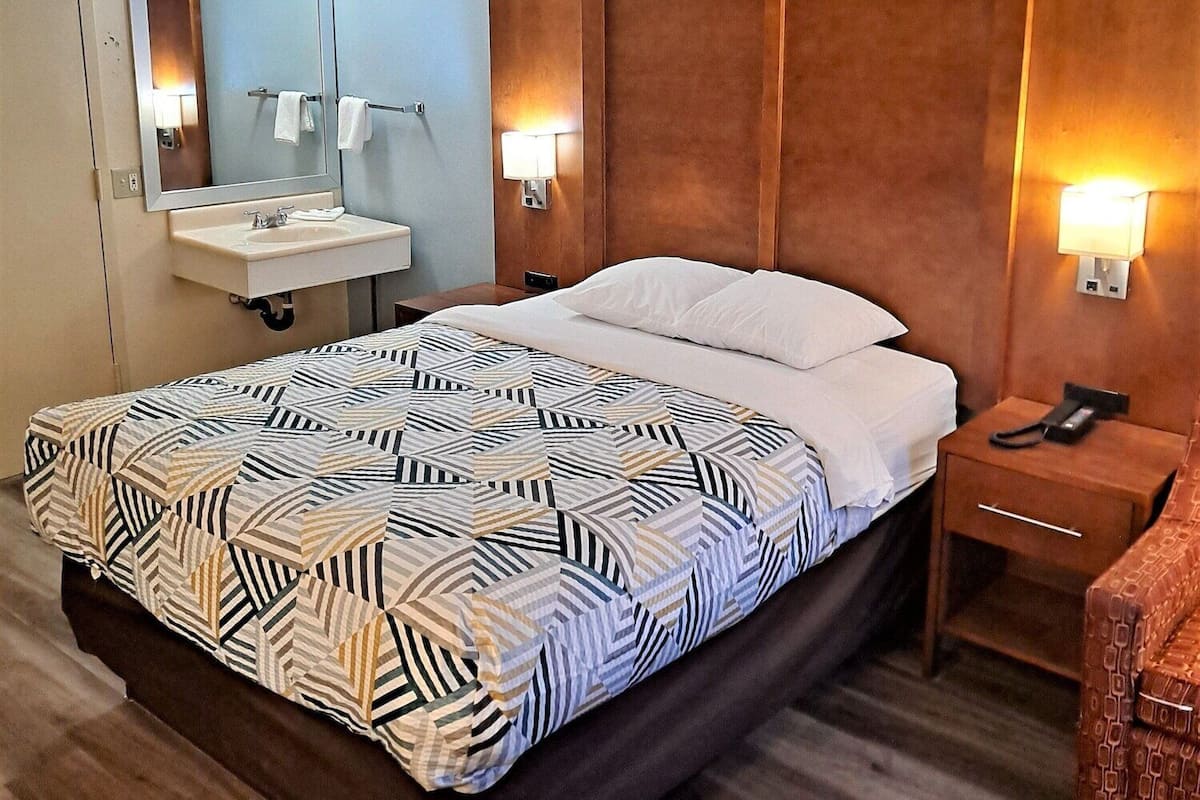 Ein Hotelzimmer mit einem Bett, einem Nachttisch mit Lampe, einem Spiegel und einem Badezimmer inklusive Waschbecken und Handtuchhalter.