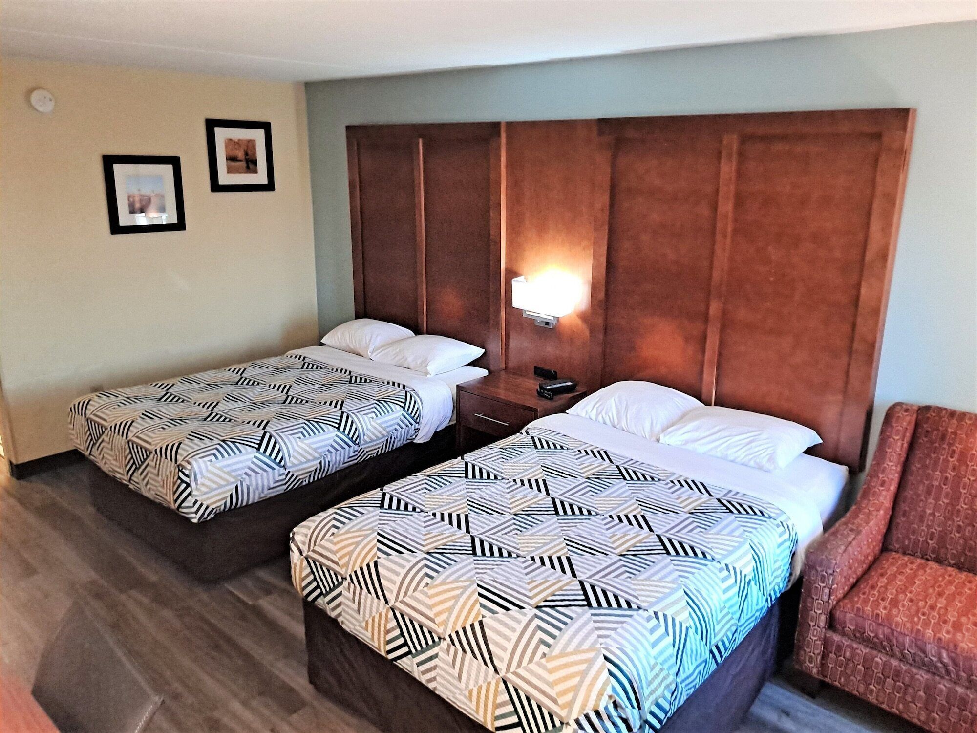 A hotel room with two beds, a nightstand, a chair, and a wall-mounted light.