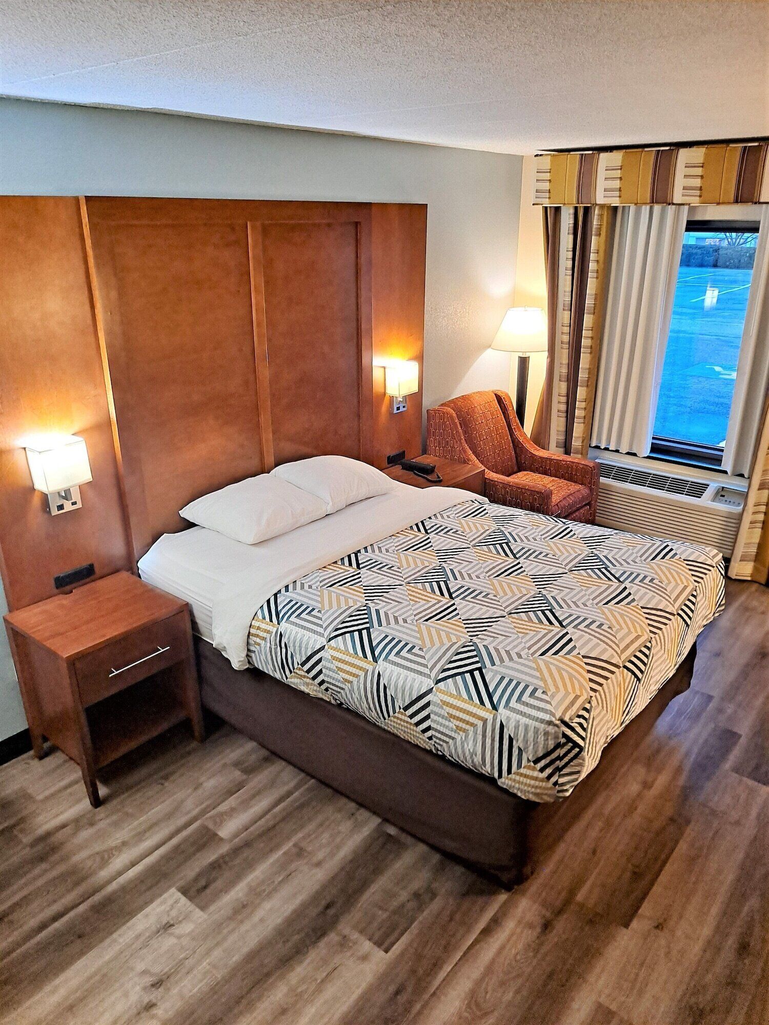Une chambre d’hôtel avec un grand lit, une table de chevet en bois, une chaise, une fenêtre avec des rideaux et une lampe fixée au mur.