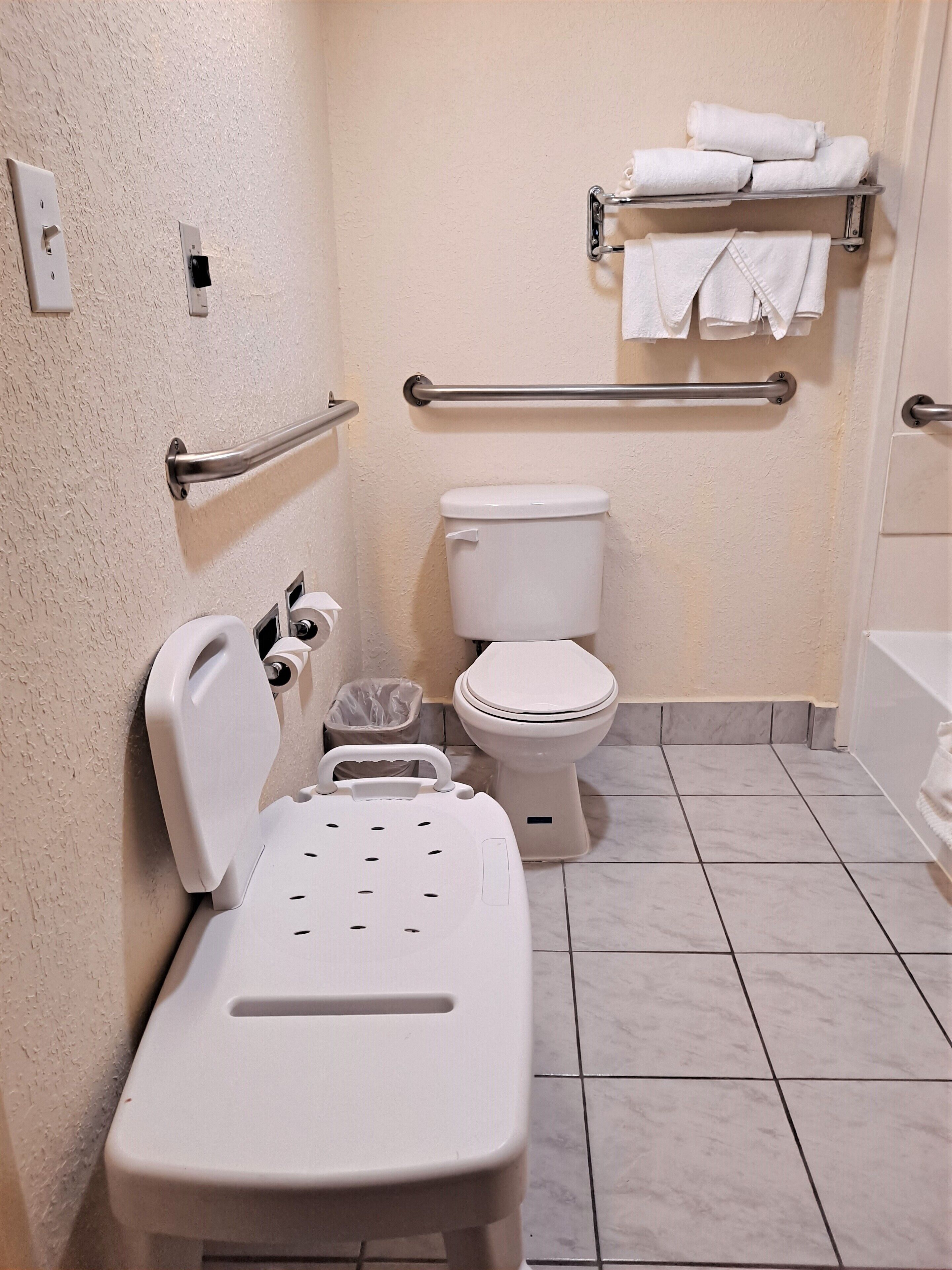 A bathroom with a toilet, grab bars, a trash bin, and a shower chair.