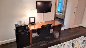 A hotel room with a wooden desk, a television mounted on the wall, a microwave, a trash can, and a patterned bedspread. - Motel 6 Catonsville, MD – Baltimore West (Catonsville)