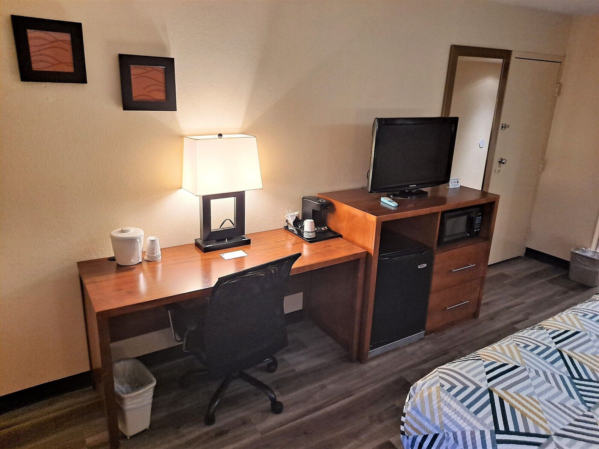 Una habitación de hotel con un escritorio de madera, una silla de oficina negra, un televisor, un microondas, una cafetera y un cubo de basura.