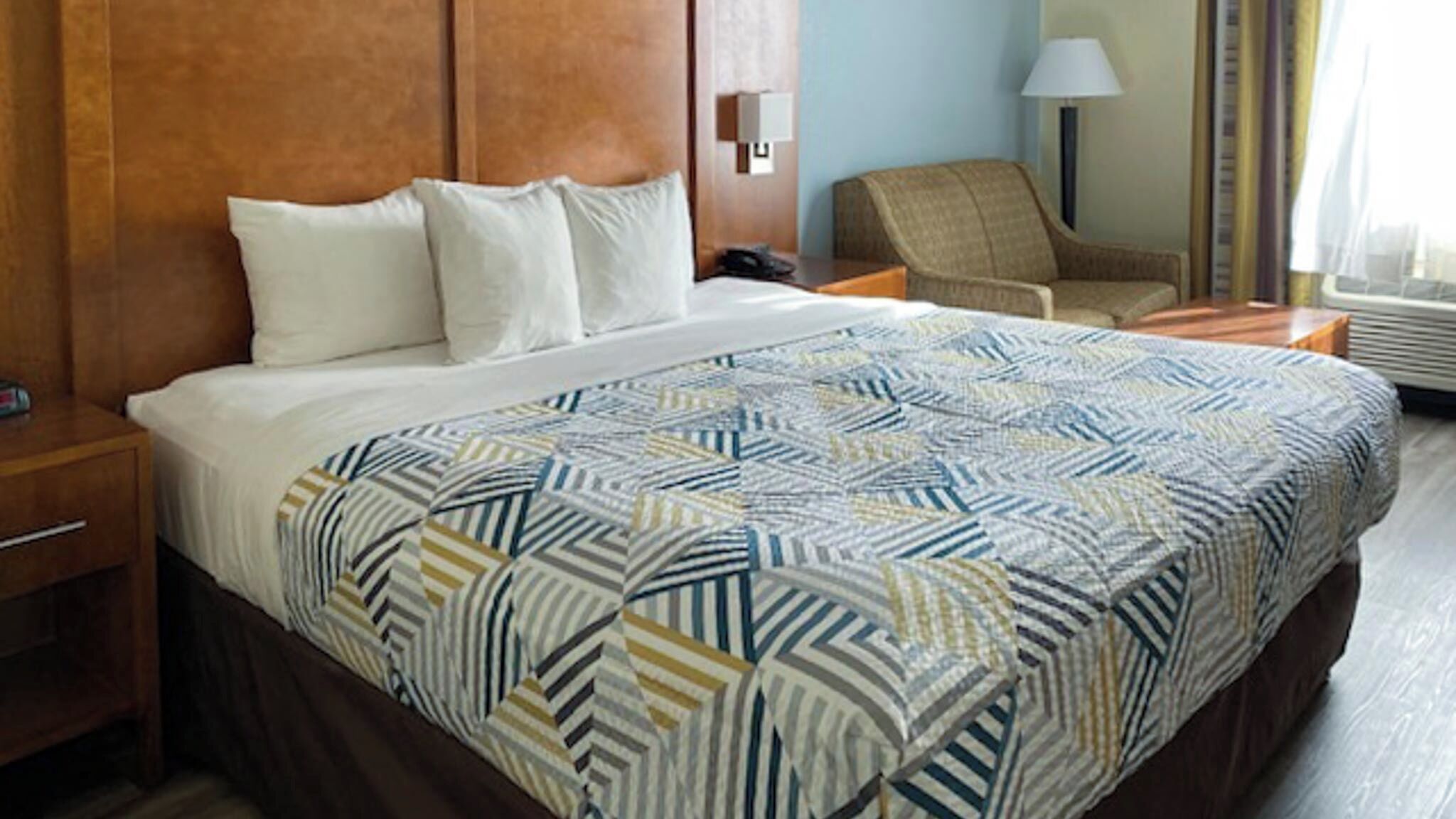 A hotel room with a large bed, a chair, a nightstand, a lamp, and a wall-mounted phone.