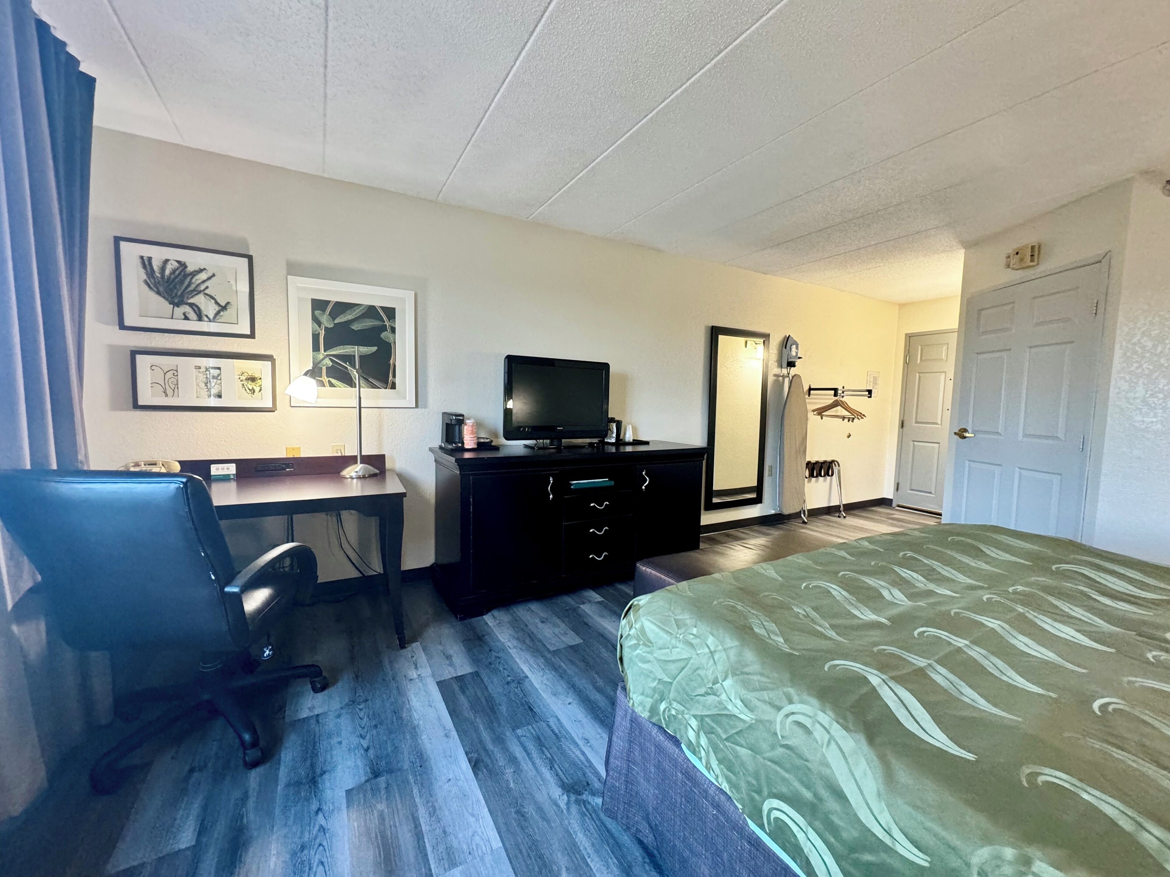 Quarto de hotel com cama, escrivaninha, cadeira, televisão e espelho.