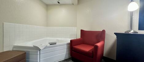 Un baño con una bañera blanca, una silla roja, un banco marrón y un escritorio con una lámpara.
