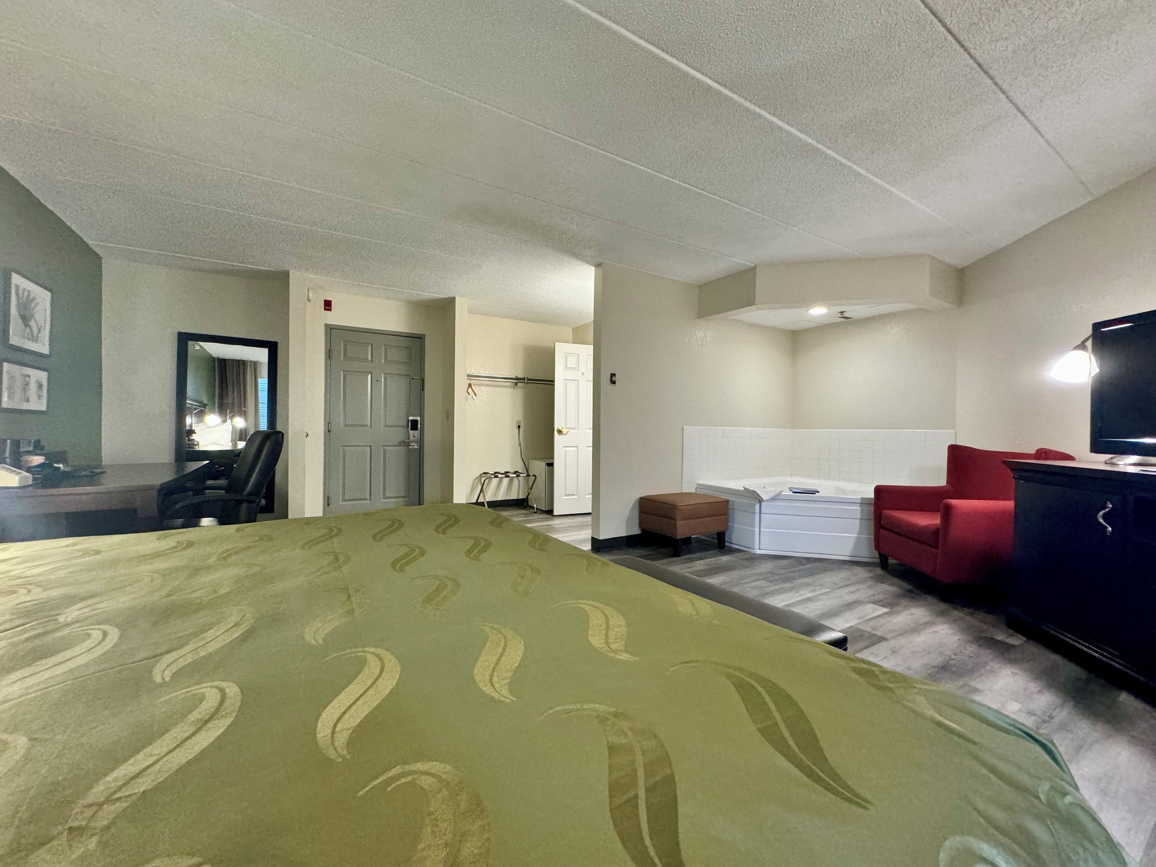 A hotel room with a bed, a desk, a chair, a red armchair, a TV, and a bathroom with a bathtub.