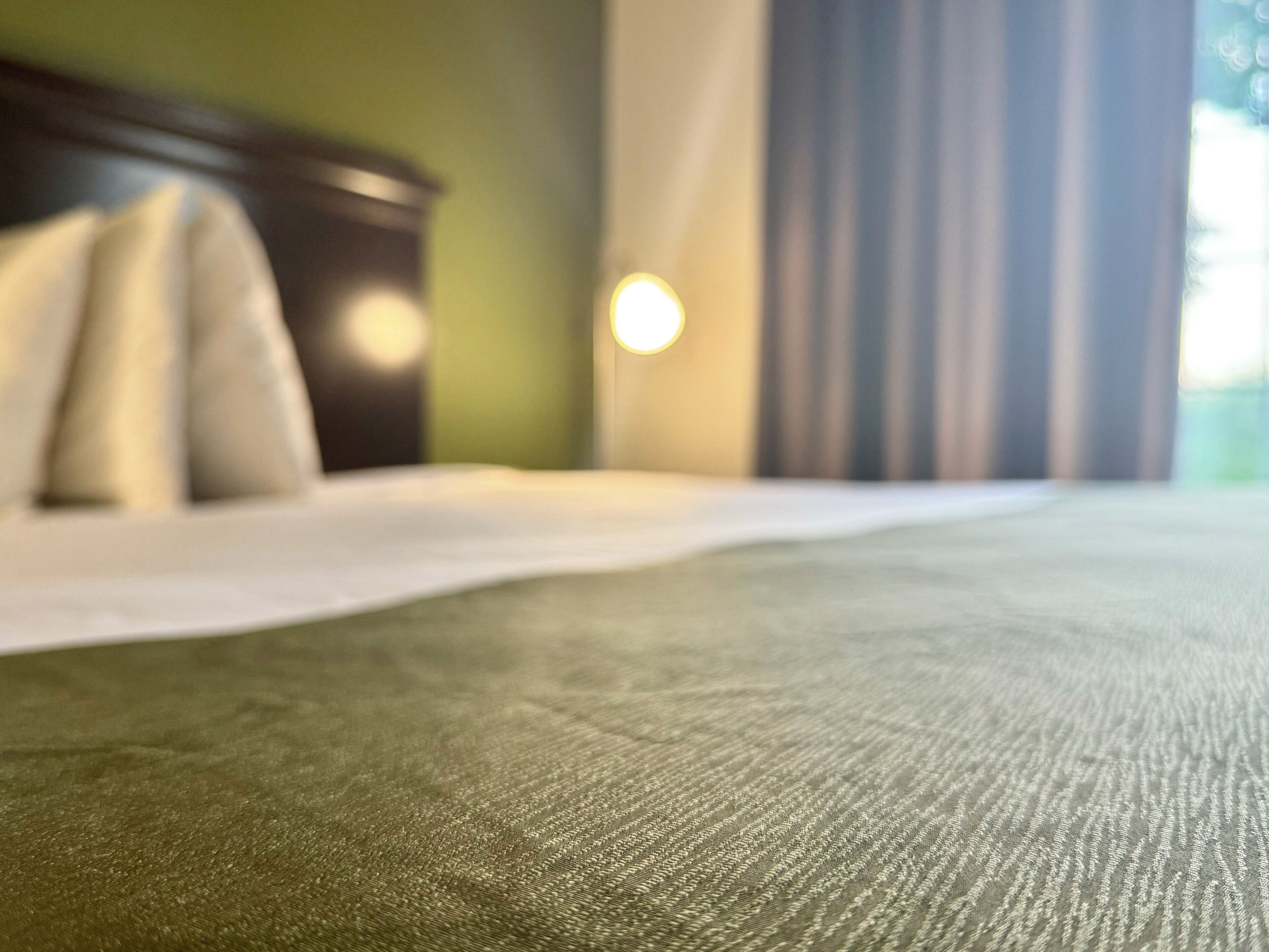 Primer plano de una cama con cubrecama verde, cabecero de madera y una ventana con cortinas.