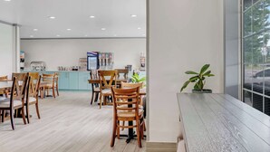 Un espace repas moderne avec des chaises et des tables en bois, une cuisine en arrière-plan et une grande fenêtre donnant sur l’extérieur.