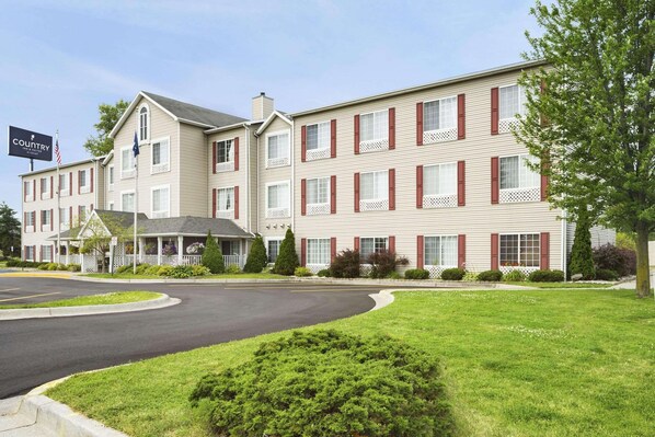 A multi-story hotel with a beige facade and red shutters, featuring a landscaped front yard and a parking area.