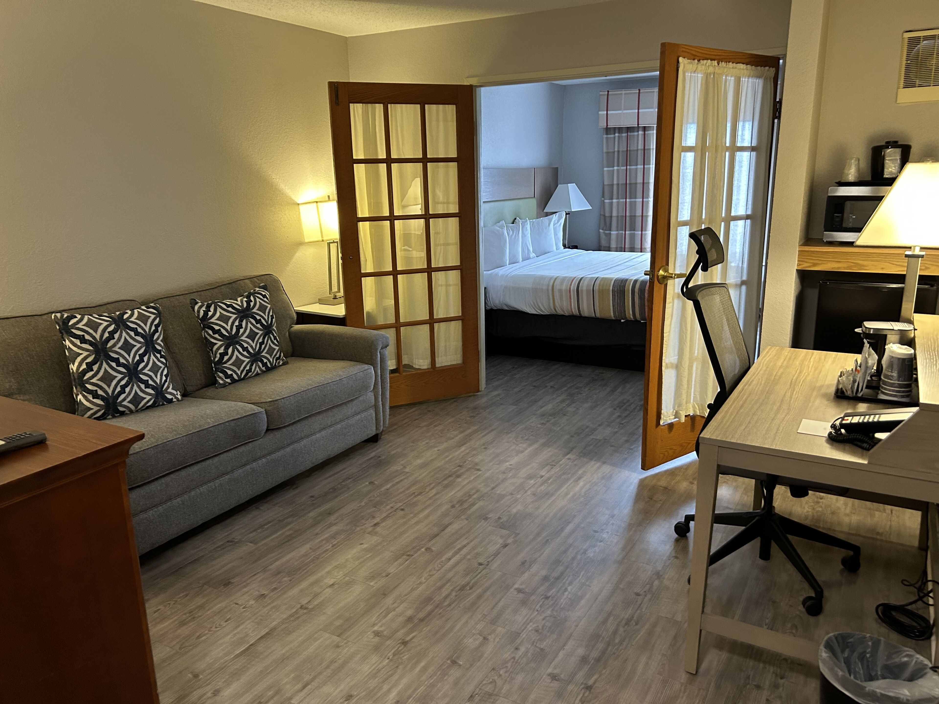 A hotel room with a grey sofa, a wooden desk, a bed with a striped bedspread, and a wooden floor.