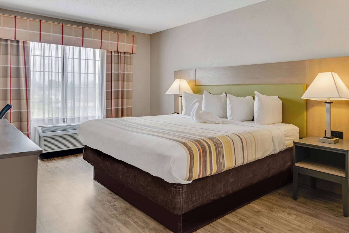 A neatly made bed with white linens and a striped blanket, a bedside table with a lamp, and a window with curtains.