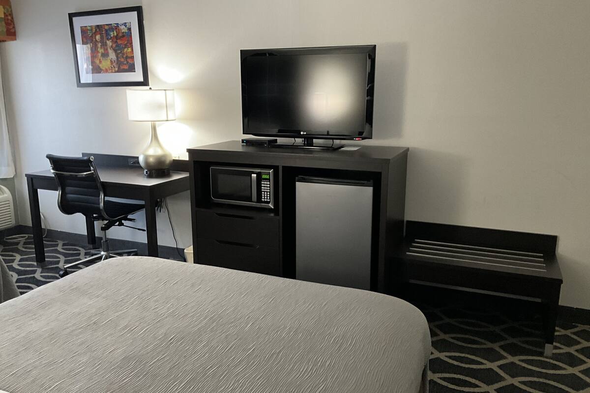 Ein Hotelzimmer mit einem Bett, einem Schreibtisch mit Computer und Fernseher, einer Mikrowelle, einer Lampe und einer Bank.