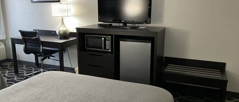 Ett hotellrum med en säng, ett skrivbord med en dator och en tv, en mikrovågsugn, en lampa och en bänk.