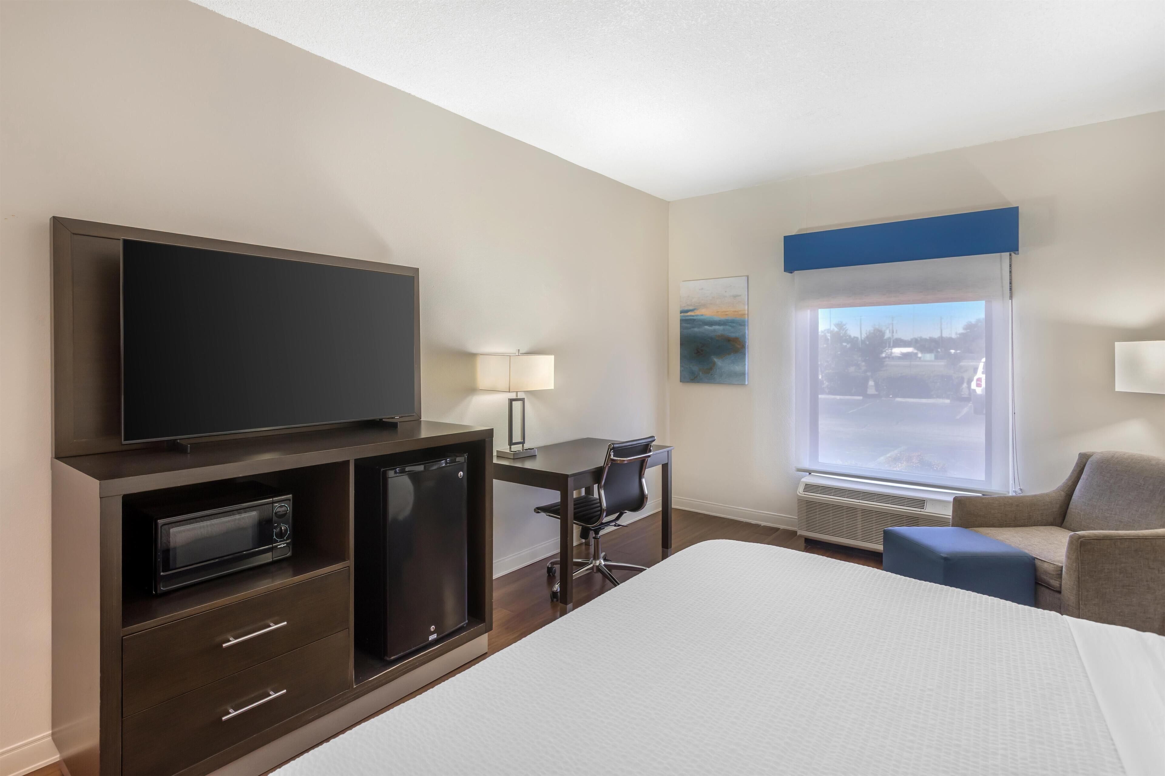 A hotel room with a bed, a desk, a television, a microwave, a chair, and a window with a view of the outside.