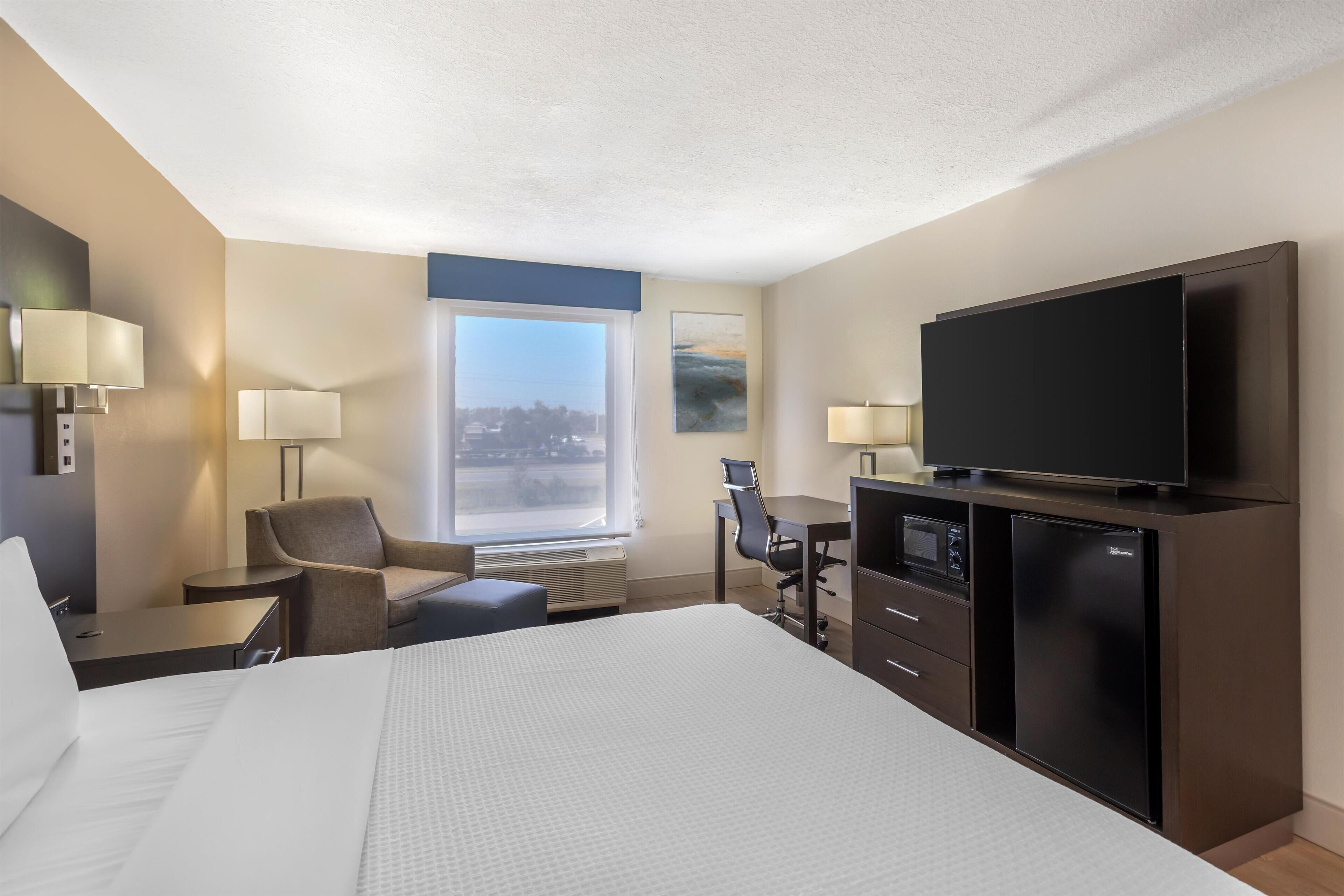 A hotel room with a large bed, a desk, a chair, a television, and a refrigerator.