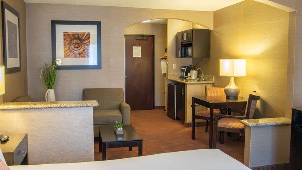 A hotel room with a bed, a desk, a chair, a small table, a lamp, a plant, a framed picture, a kitchenette, and a door. - Country Inn & Suites by Radisson, Garden City, KS (Garden City)