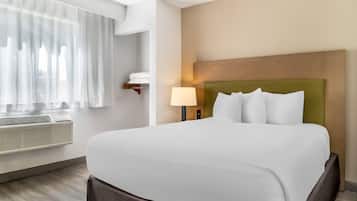 A hotel room with a large bed, a bedside lamp, a window with sheer curtains, and a wall-mounted air conditioner.