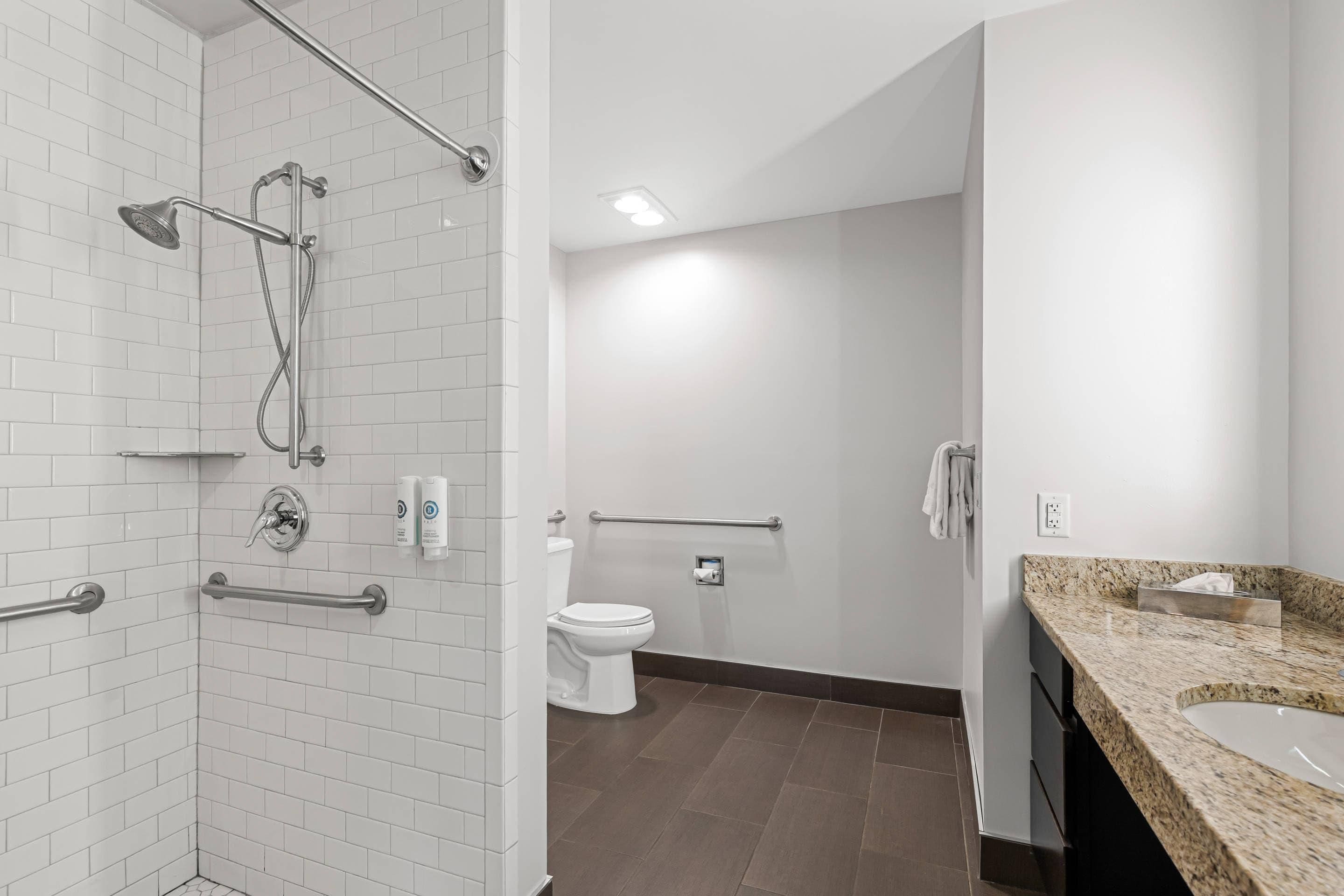 A modern bathroom with white subway tiles, a shower with a handheld showerhead, a toilet, and a granite countertop with a sink.