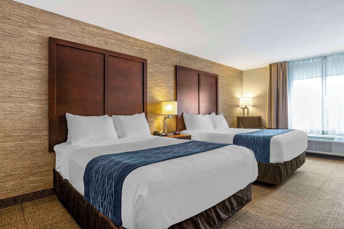 A hotel room with two beds, a large window with curtains, a wall-mounted lamp, and a textured wall.