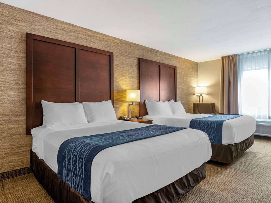 Ett hotellrum med två sängar, ett stort fönster med gardiner, en vägglampa och en vägg med en intressant yta.