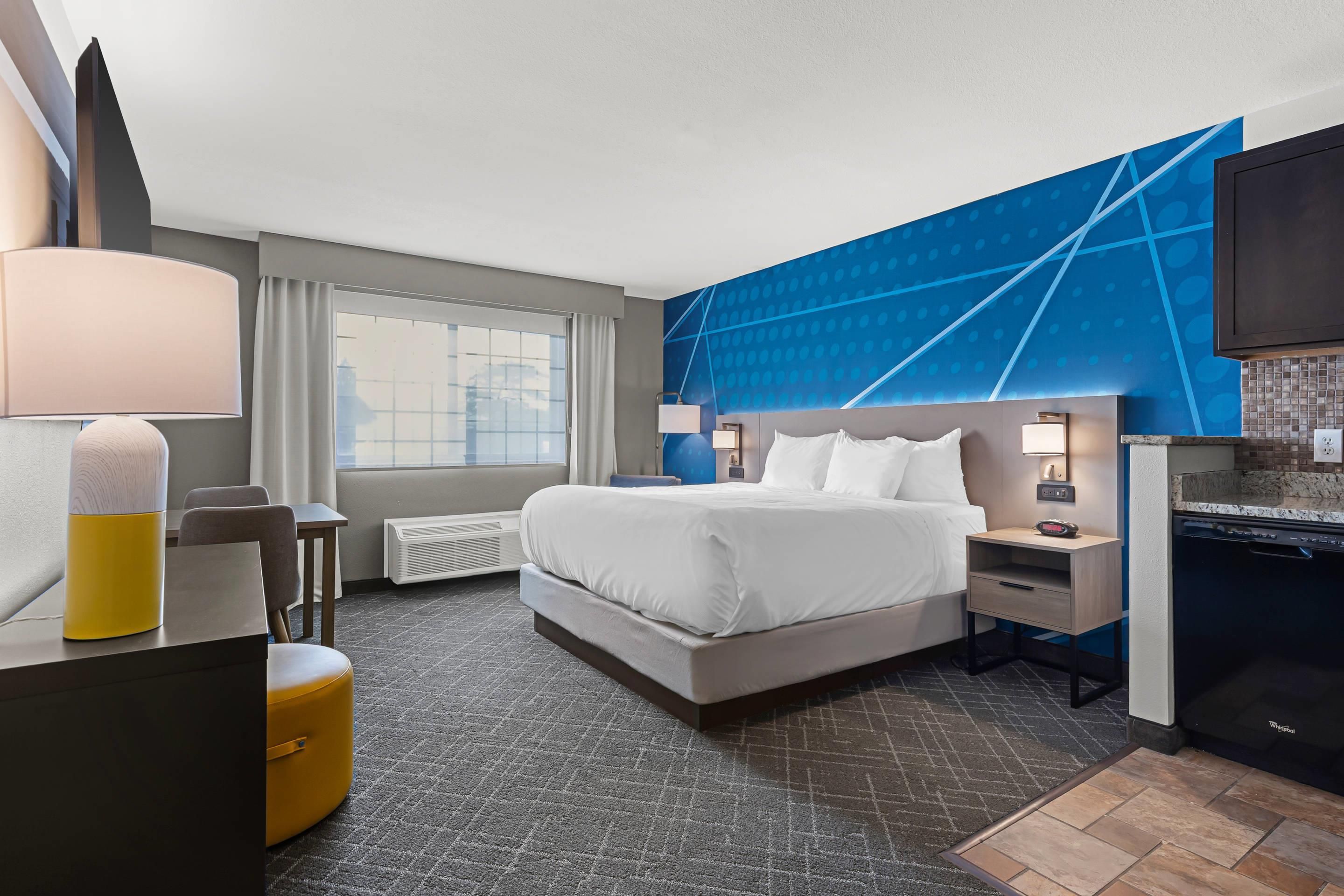 A hotel room with a bed, bedside tables, a desk with a chair, a large window, and a wall-mounted TV.