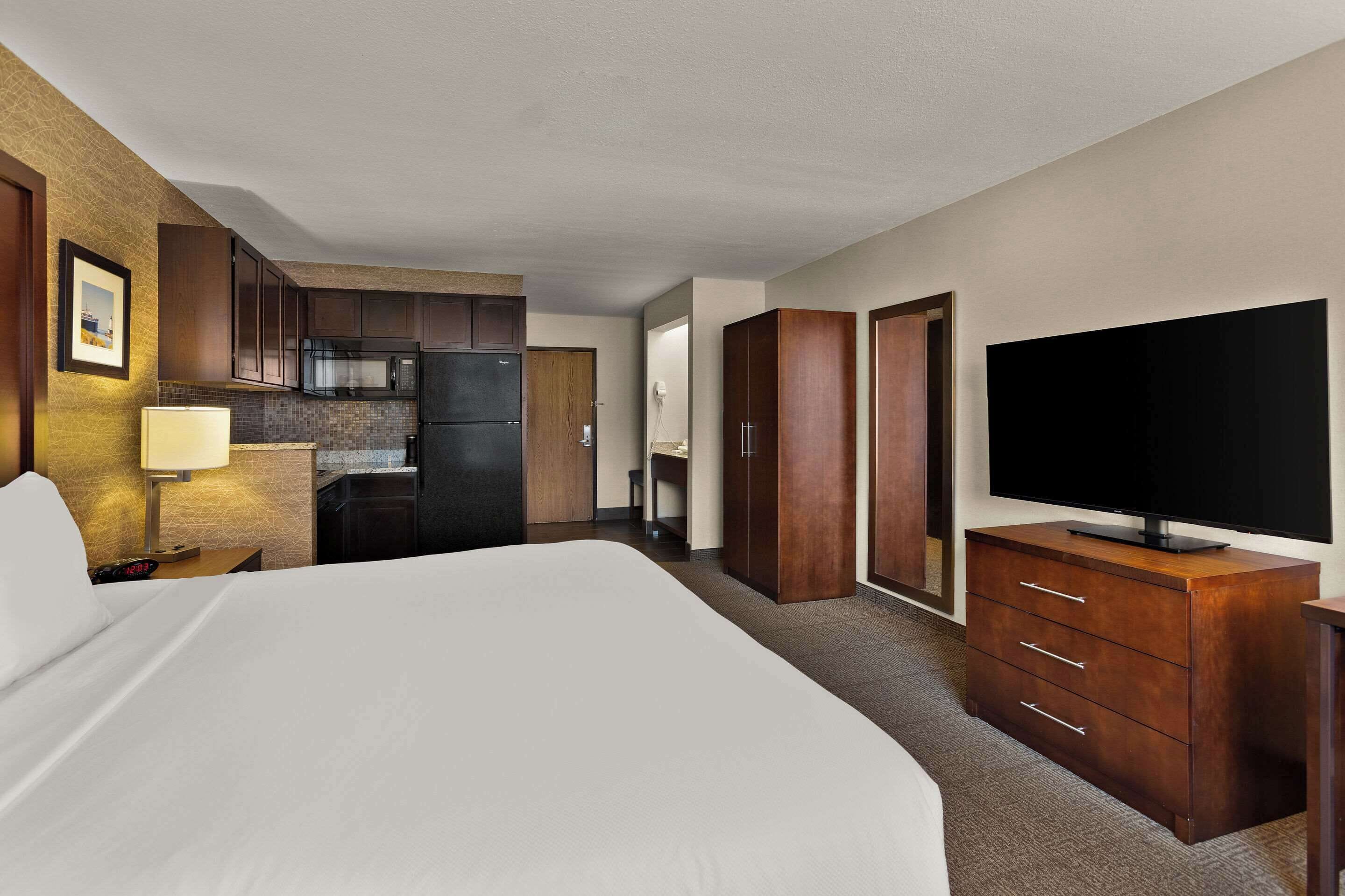 A hotel room with a bed, a TV on a wooden dresser, a kitchen area, and a bathroom.