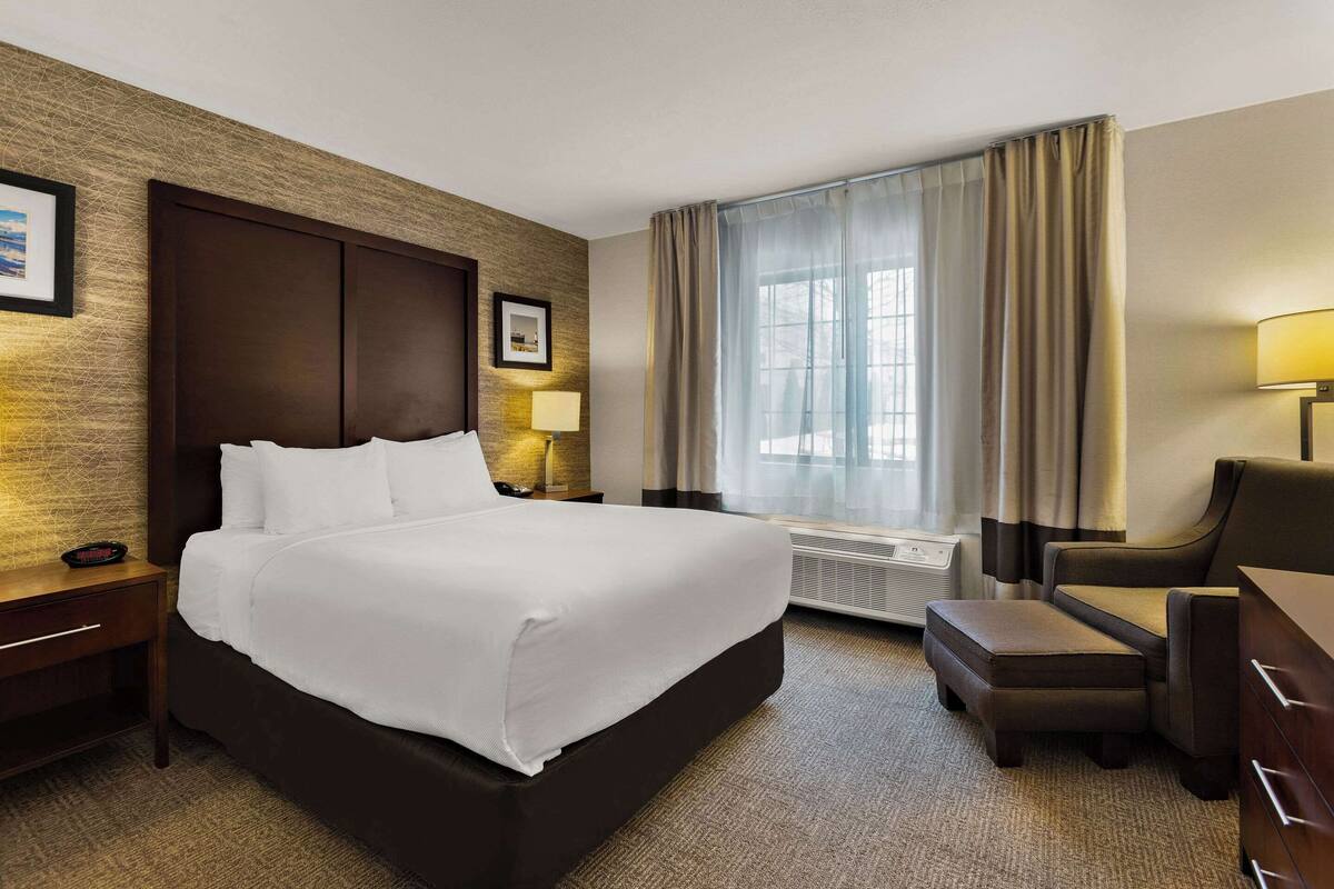 A hotel room with a large bed, a desk, a chair, a window with curtains, and a wall-mounted air conditioner.