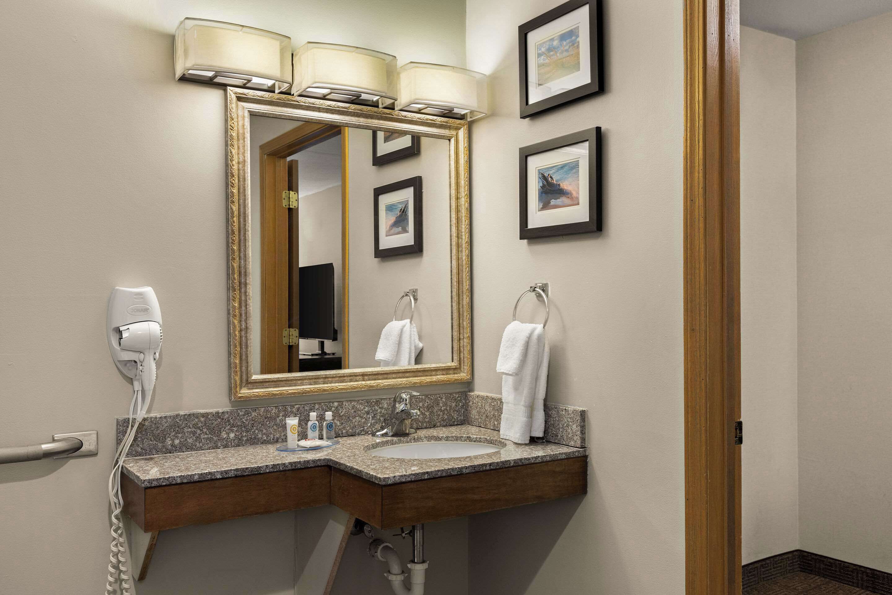 A hotel bathroom with a granite countertop sink, a large mirror with a gold frame, a hair dryer, and framed pictures on the wall.
